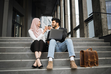 Happy muslim man and woman toasting with cups of coffee while sitting together on outdoor stairs....