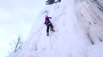 Low angle shot of an alpinist woman with ice climbing equipment, axe and climbing ropes, climbing  at a frozen waterfall, view from below