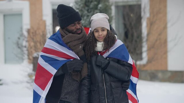Portrait of happy African American man and girl wrapped in British flag posing outdoors on winter day. Medium shot of smiling father and daughter hugging looking at camera