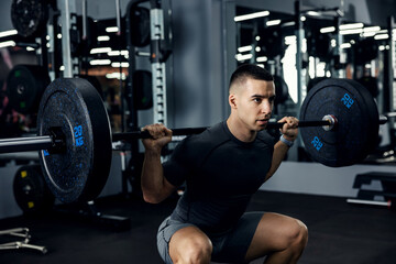 Close up side portrait of a male fitness trainer in grey sportswear doing squats using a barbell in...