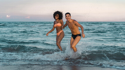 Full length portrait of Interracial young couple having fun relaxing on the beach holding hands on the water. Summertime fun concept.