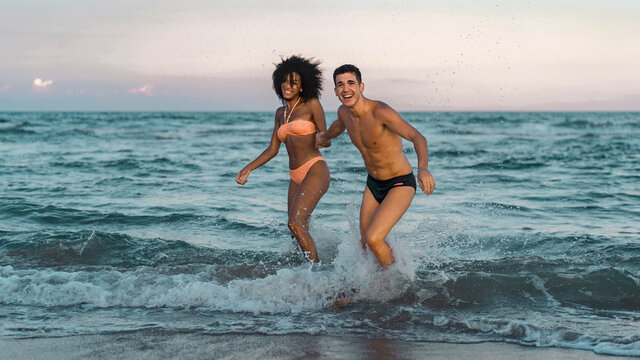 Full length portrait of Interracial young couple having fun relaxing on the beach holding hands on the water. Summertime fun concept. - Powered by Adobe