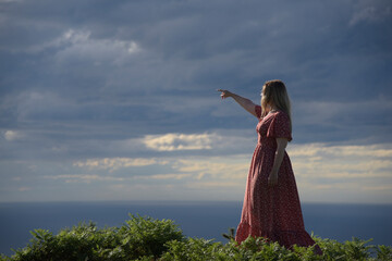 Mujer rubia con vestido rojo, en el monte señalando al sol con unas nubes oscuras detrás. 