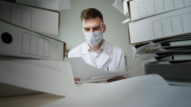 Tired young doctor wearing protective face mask and gown working with pile of folders, medical student parsing documents having headache, exhausted male nurse, fatigue, lots of work during covid-19. 