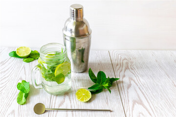 Fresh iced cold mojito lemonade summer drink, cocktail with sliced lime, bright green mint leaves, shaker and frozen ice cubes in a transparent cup, mason jar on wooden table