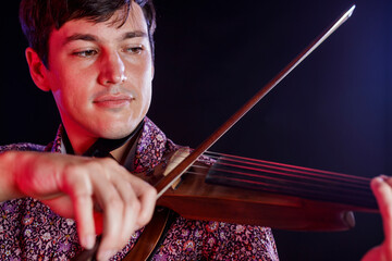 Close-up of a young man playing a 5-string electric violin. Dark background. Blue and red lights....