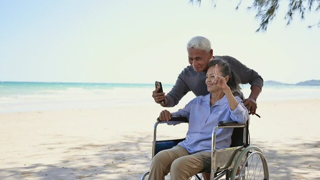 Asian senior couple tourist smile and pose by hands symbol to camera video call and make selfie by smartphone at beach. Couple elderly retire rest and relax wife is disabled and sitting on wheelchair.