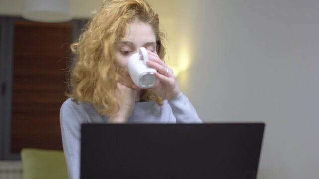 Cute girl sitting on a table in living room, infront of a laptop computer, drinking coffee. Portrait of a womam working from home. Camera moving right.