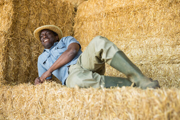 Cheerful adult african american farm worker in straw hats resting on haystack during break from work