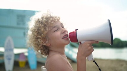 Young pretty curly boy sitting on high white chair with lifeline and posing with megaphone on the beach against blue lifeguard tower. 