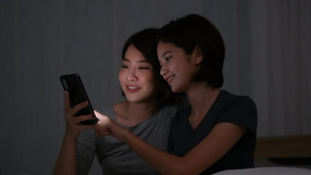 Asian couple women use mobile phone and look enjoy together in bedroom at night. Concept of good relationship between friends during stay at home.