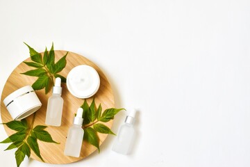 White bottles with pipettes, moisturizer cream jars on a round wooden cut with green leaves on a white background. Set of eco natural cosmetics. Copy space. Bioorganic cosmetic product.