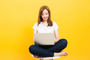 Asian happy portrait beautiful cute young woman teen smiling sitting crossed legs on a chair with laptop computer looking to computer isolated, studio shot on yellow background with copy space