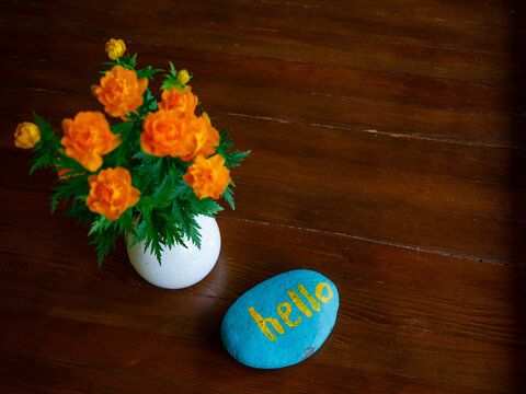 vase with orange spring summer globe flowers Trollius asiaticus and blue stone with word hello on wooden table background. concept hello background, horizontal, top view, copy space