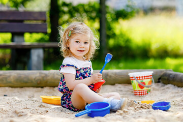 Cute toddler girl playing in sand on outdoor playground. Beautiful baby in red trousers having fun on sunny warm summer day. Child with colorful sand toys. Healthy active baby outdoors plays games