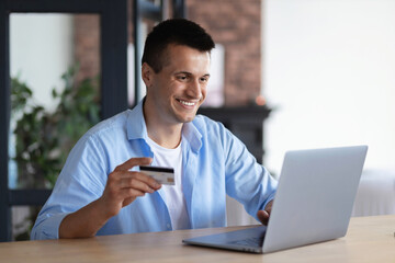 Fototapeta na wymiar Happy smiling young male or student with a credit card using laptop for profitable purchases in the Internet sitting at home, online shopping