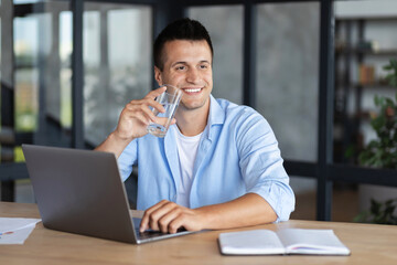 Young successful businessman man drinking clean water sitting at the office table. Smiling freelancer using laptop for working,  holding glass of fresh water in his hand