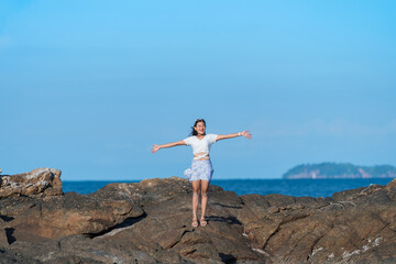 happy Asian woman in swimwear standing on rock near blue sea with arms open, relax on vacation at rock beach with blue sky, freedom nature travel concept