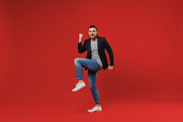 Full length young businessman latin man 20s with long hair ponytail wearing black striped jacket grey shirt look camera do winner gesture with raised up leg isolated on red background studio portrait