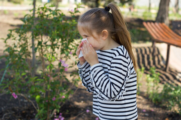 A girl with blond hair in a striped T-shirt sneezes near a bush with flowers in the park, covering her nose with a napkin. Seasonal bloom allergy. Selective focus.