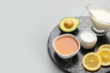 Bowl with sweet honey, avocado, lemon slices, jug of milk and cosmetic product on grey background