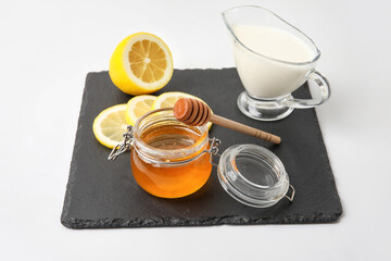 Glass jar with sweet honey, lemon slices and jug of milk on slate board against white background