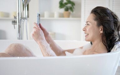 Attractive young mixed race asian woman making video call using smartphone lying in bathtub. Millennial girl in bath with mobile phone having webcam video conversation enjoy daily bodycare in bathroom - Powered by Adobe