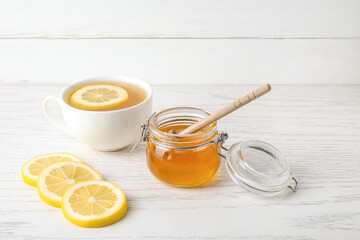 Glass jar with sweet honey, cup of tea and lemon slices on light wooden background