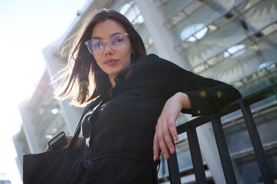 Business woman portrait in city streets at sunset. Sexy brunette wearing black dress, glasses and black leather bag