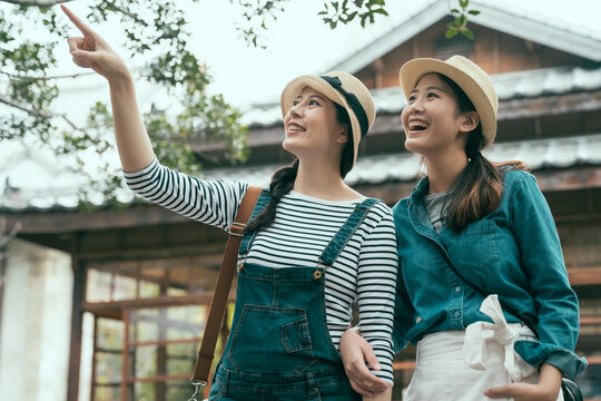 happy two young asian women friends walking outdoors standing by japanese wooden house. ladies looking aside while pointing finger to sky. cheerful female travelers smiling share beautiful scenery