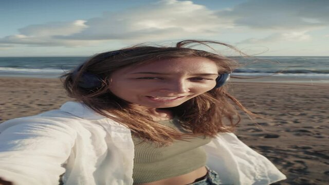 Vertical handheld POV of joyous Caucasian woman wearing over-ear headset, standing on sandy ocean coast, filming video on selfie camera, talking and smiling