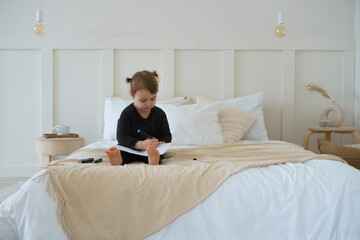 a little girl in black home clothes is sitting on the bed and drawing with felt-tip pens