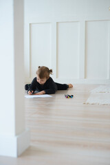 a little girl in black home clothes and barefoot lies on the floor and draws with felt-tip pens on a piece of paper