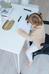 маленькая девочка в светлой домашней одежде сидит за белым столом рисует вид сверху