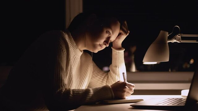 Tired female student. Online school. Distance education. Overload information. Exhausted woman almost sleeping looking laptop writing notebook light table lamp dark night home interior.