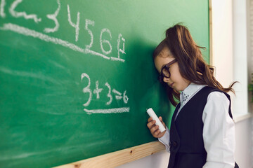Exhausted girl in eyeglasses leaning on green chalkboard and doing sums during math lesson in school