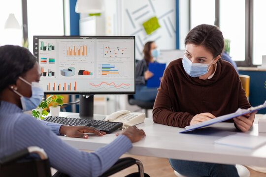 Immobilized handicapped invalid african entrepreneur working with colleague in new normal business office during covid19 pandemic wearing face masks, checking graphs calculating profit on computer.