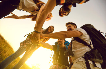Fototapeta na wymiar From below of a team of happy young friends joining hands ready for great travel adventure. Low angle of group of tourists with backpacks stacking hands in summer field while hiking in countryside