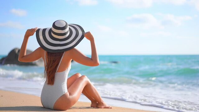 A young woman in a bathing suit and floppy sun hat sits on a sandy beach raising her arms in welcome to the incoming surf. Title space