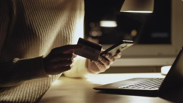 Mobile payment. Online shopping. Banking transfer. Unrecognizable woman holding plastic card typing smartphone verification number dark shadow light lamp room interior.