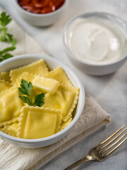 Ravioli in a white bowl with a lump of butter and a green parsley leaf on top. Nearby is a white bowl of sour cream. White background. Close-up. Careful viewing.