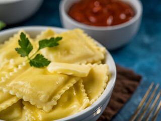 Table set for dinner. Close-up of a bowl of ravioli. In the background is a bowl of tomato sauce. Brown napkin, blue background. Careful viewing. Close-up.High angle view.