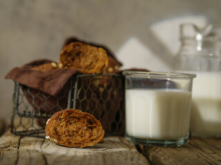 On a wooden table, chocolate chip cookies in a basket. Milk in a glass glass. In the background is a jug of milk. White and golden tones. Close-up. Color image.