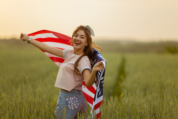 young happy beautiful  girl in pink top and jeans shorts with national American flag  in green wheat field at sunset.4th of July celebrating