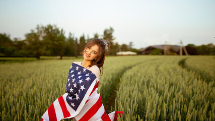young happy beautiful  American  girl in pink top and jeans shorts covering national USA flag  in green wheat field at sunset. celebrating 4th of July 
