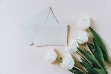 Still life scene with white tulips bouquet on beige background and blank business, greeting card, invitation mockup with envelope.