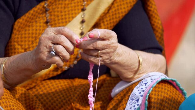 An Elderly Indian Lady Knitting Woolen Clothes With The Help Of A Crochet Hook. Closeup Shot Of An Old Woman Hands Knitting A Scarf With Pink Colored Yarn During Leisure Time - Hobby Concept