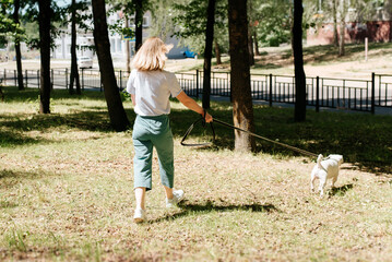 Young woman in stylish clothes walking in the park with her dog, rear view. Blonde girl and jack russell terrier on a leash for a walk outdoors, sunny summer day.