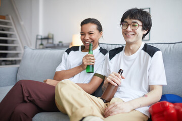 Portrait of two cheerful young people watching TV and drinking beer at home, copy space