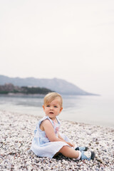 Fototapeta na wymiar Little girl in a dress sits on a pebble beach by the sea